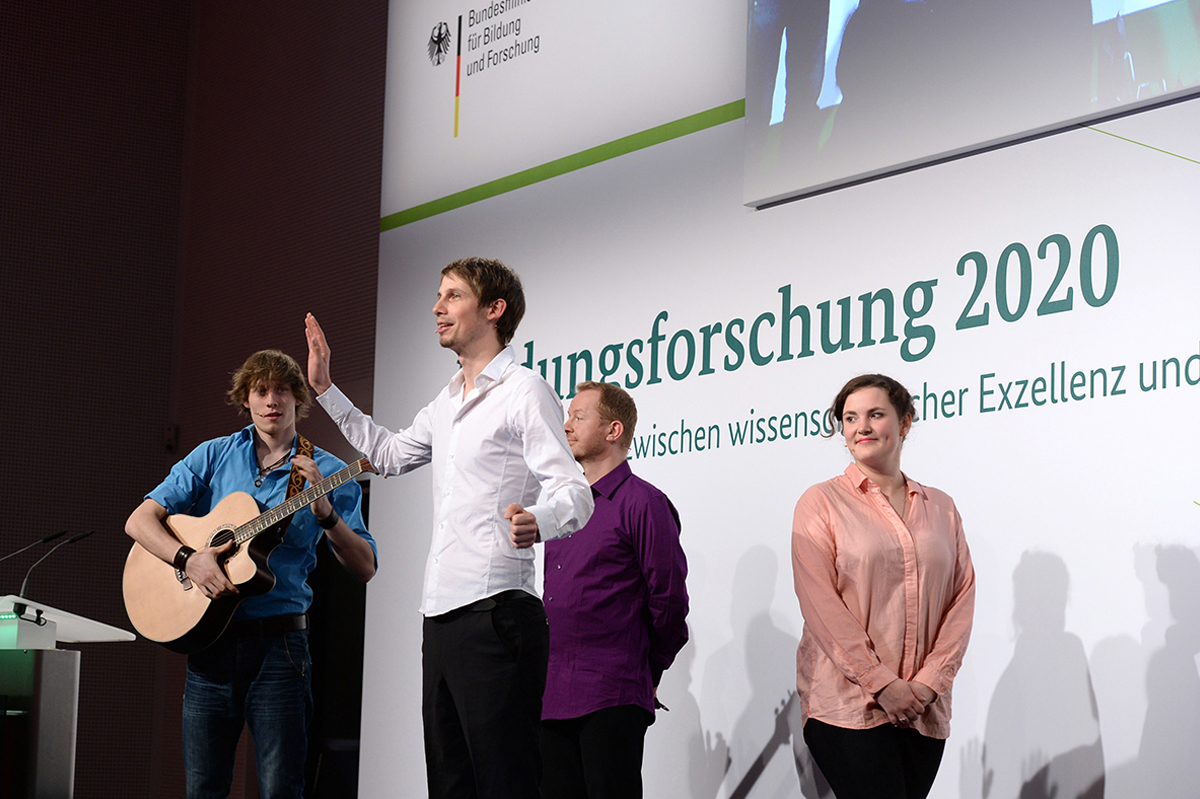 Vier Personen stehen auf der Bühne, eine Person davon hat eine Gitarre über der Schulter und eine andere hebt ihre Hand