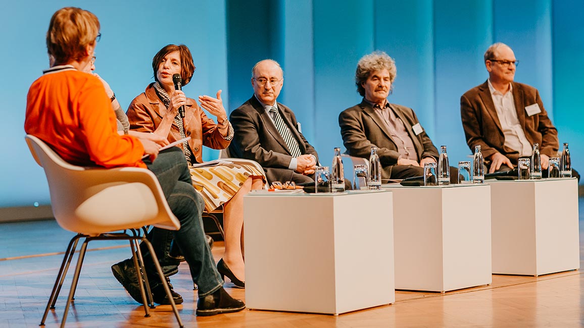 Fünf Personen sitzen bei der Podiumsdiskussion auf der Bühne, eine Person hält ein Mikrofon in der Hand