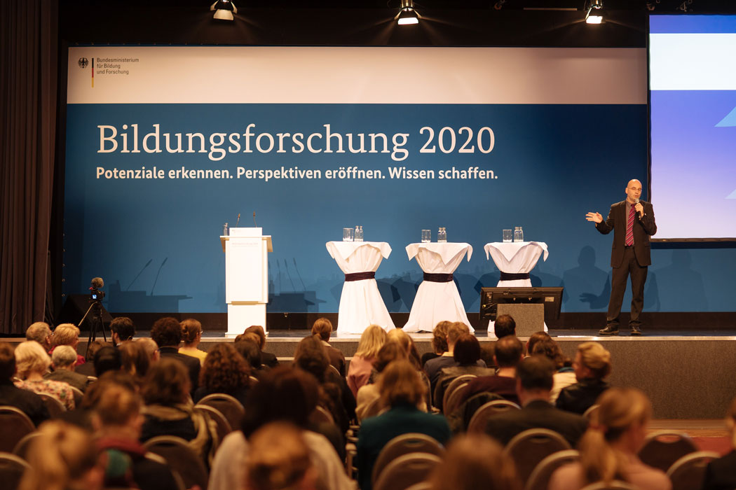 Der Moderator steht mit einem Mikrofon in der Hand auf der Bühne vor einem Plakat mit dem Titel Bildungsforschungstagung 2020, vor der Bühne ist sitzt das Publikum