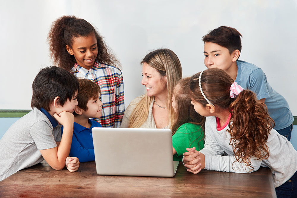 Kinder schauen gemeinsam mit einer Lehrkraft auf den Bildschirm eines Laptop